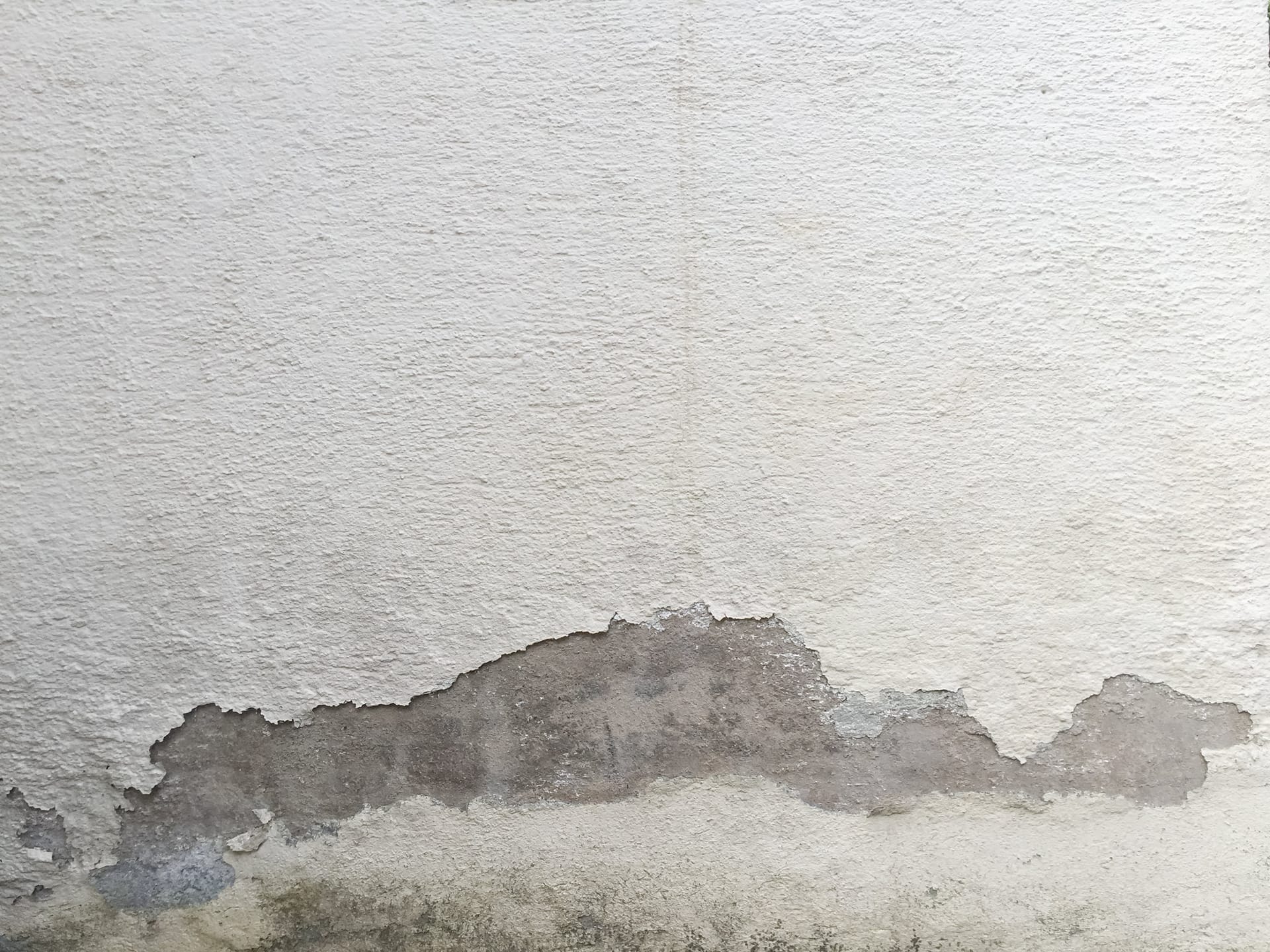 Mancha oscura de humedad ascendente en la parte baja de una pared enlucida en blanco.