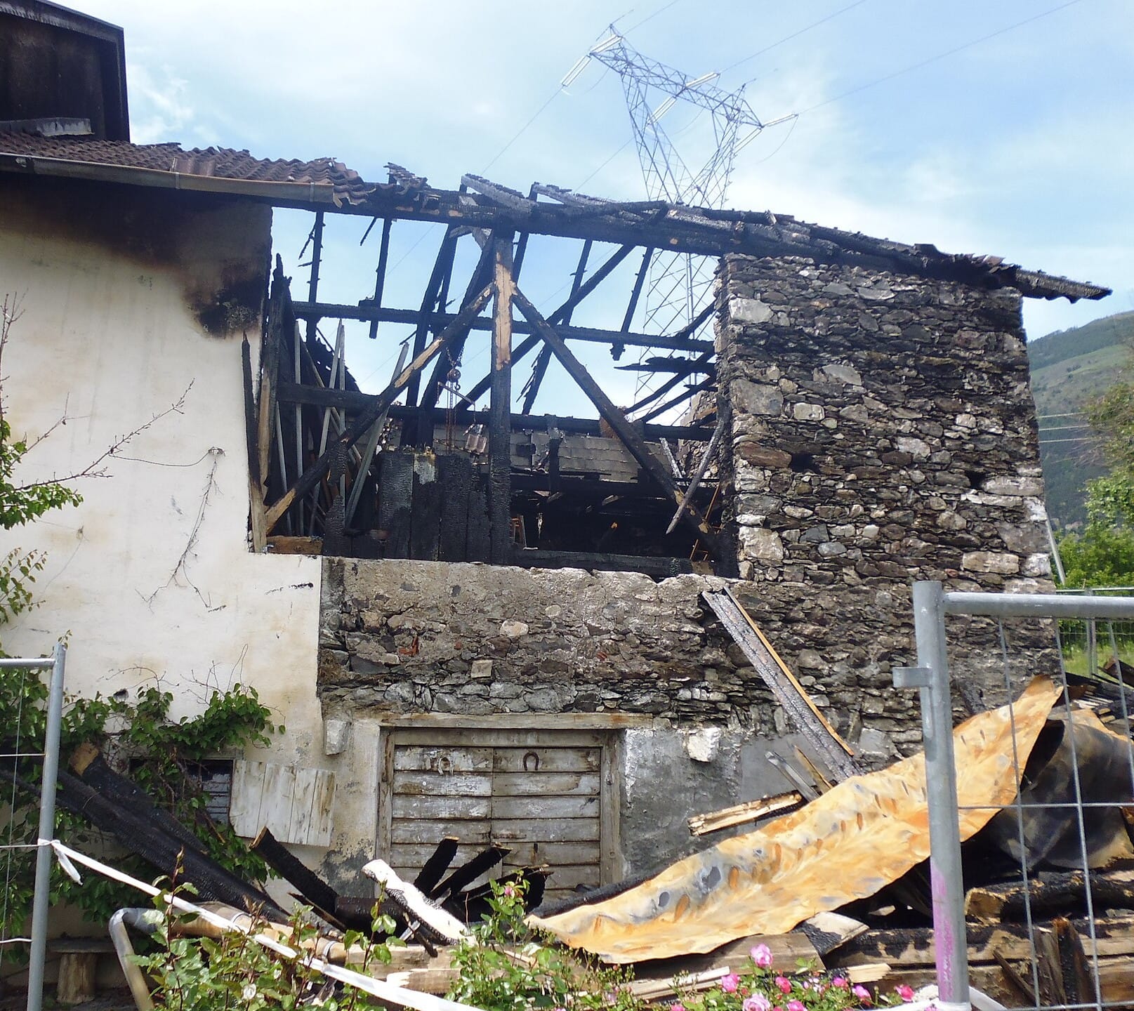 Casa rural de piedra con la cubierta de madera calcinada tras un incendio, marcas de hollín en la fachada y escombros quemados tras una valla de obra.