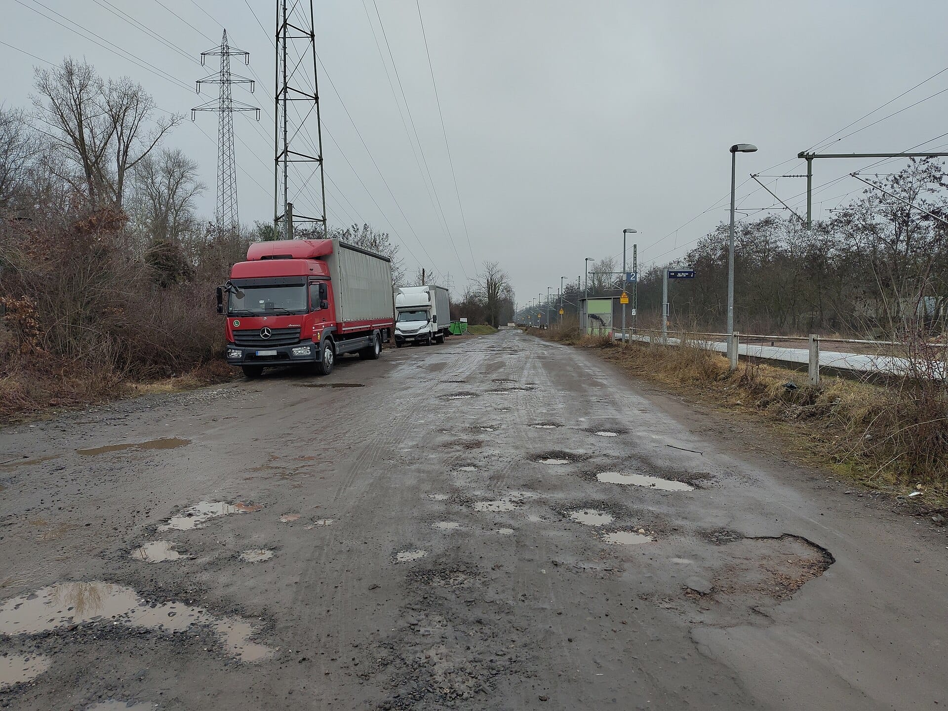 Vial de asfalto muy deteriorado con baches llenos de agua y camiones aparcados en una zona industrial.