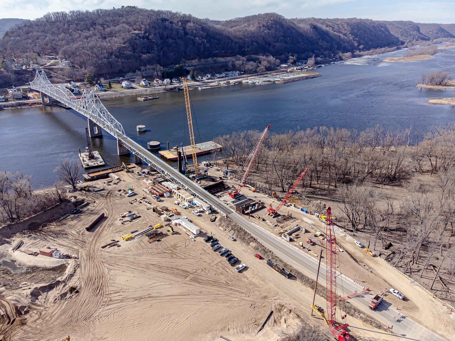 Aerial view of a new bridge under construction over a wide river, with cranes, barges and material stockpiles beside the existing bridge.