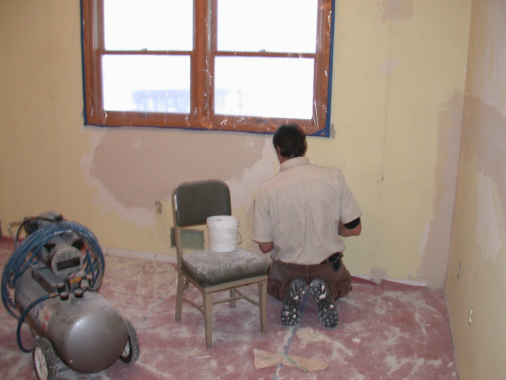 Operari d'esquena reparant una paret amb pedaços de massilla en una habitació en obres, amb el terra protegit i un compressor.